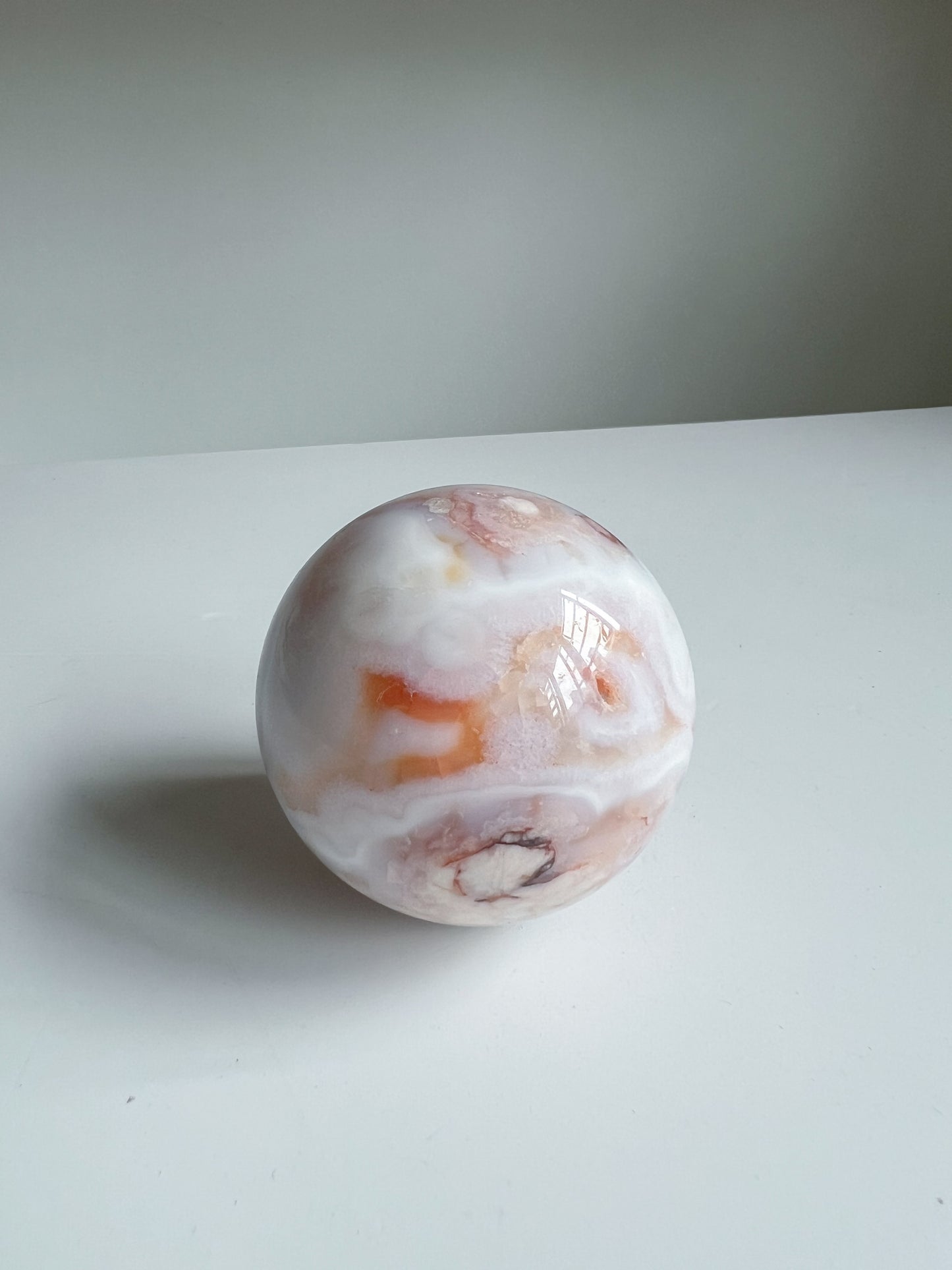 Aurora carnelian flower agate crystal sphere featuring bold orange and white banding with soft pink undertones and a dark druzy detail, placed on a white background