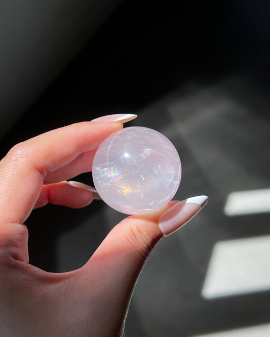 Translucent Girasol Rose Quartz sphere glowing in direct sunlight, revealing inner veils and a bright rainbow flash on its surface.