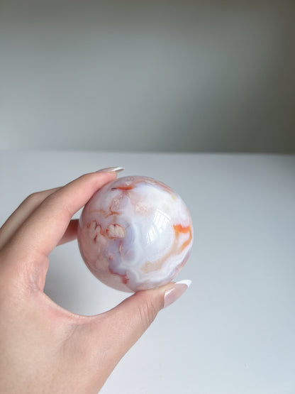 Aurora carnelian flower agate crystal sphere held in hand, showing soft pastel banding in peach, pink, red, blue, and white tones against a white background