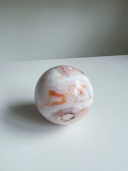 Aurora carnelian flower agate crystal sphere featuring bold orange and white banding with soft pink undertones and a dark druzy detail, placed on a white background