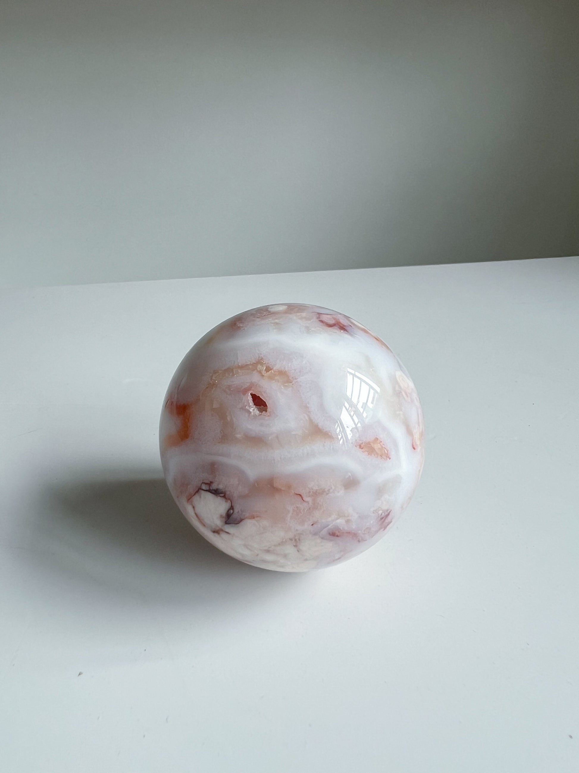 Aurora carnelian flower agate crystal sphere displaying peach and cream banding with subtle inclusions and a dark druzy accent on a white background