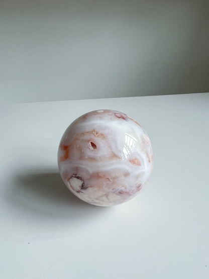 Aurora carnelian flower agate crystal sphere displaying peach and cream banding with subtle inclusions and a dark druzy accent on a white background