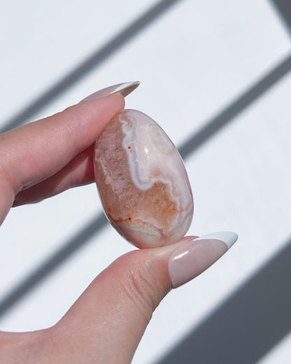 Hand holding a Carnelian Flower Agate bean with orange scalloped banding and translucent cream veils under sunlight.