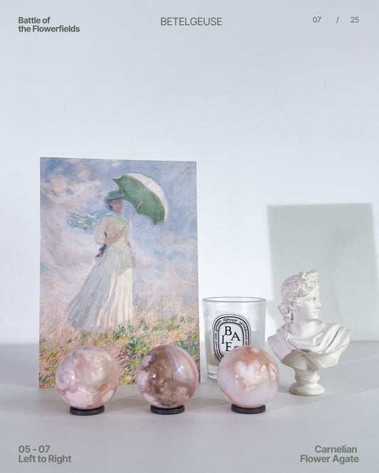Image shows three Carnelian Flower Agate spheres (05–07) arranged left to right in a still-life setup featuring a Monet-style art print, a Diptyque glass candle, and a white bust. Soft indoor lighting highlights the pastel hues and floral plumes of each sphere.