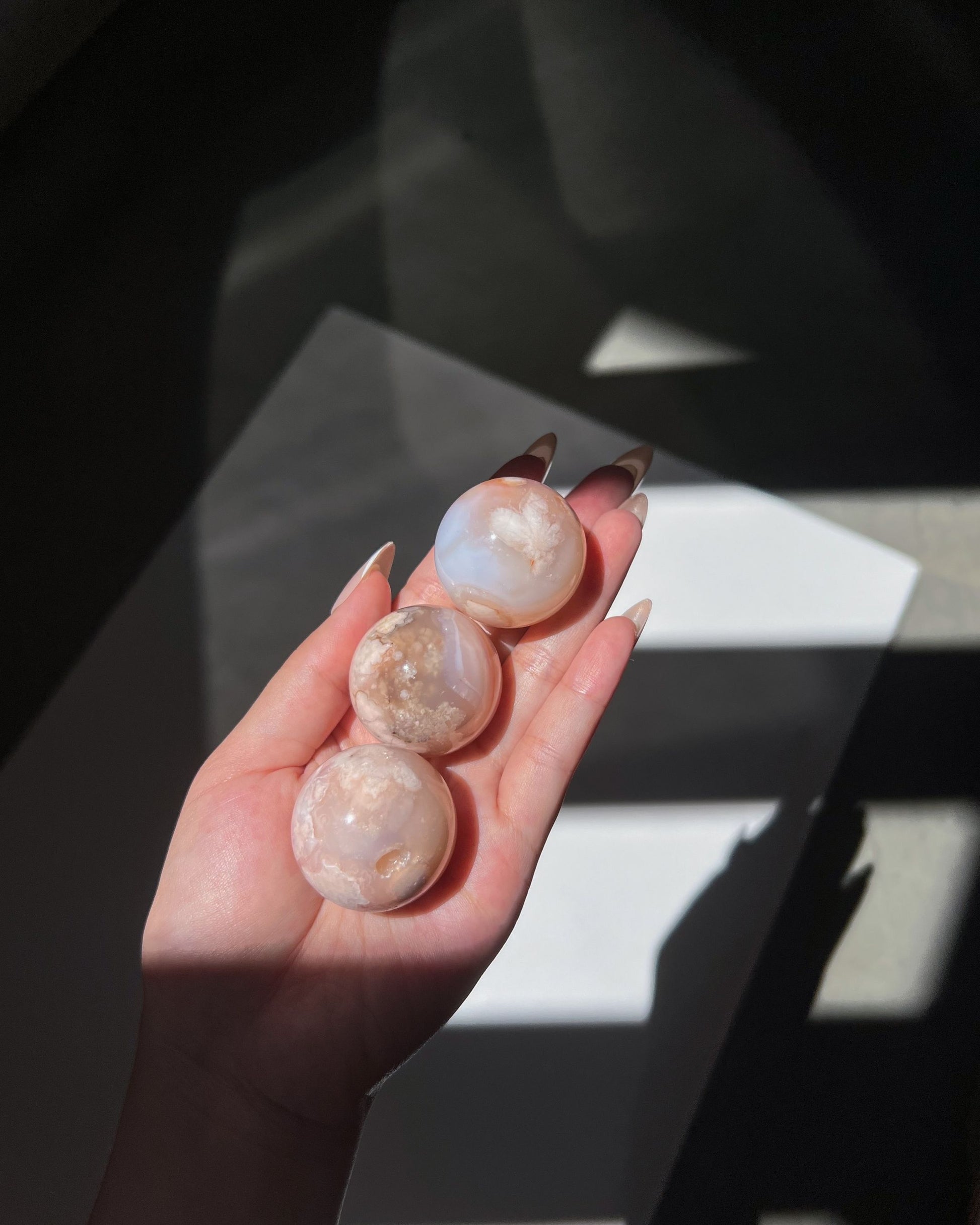 A different angle of the same hand holding three Carnelian Flower Agate spheres. Light and shadows create a dreamy contrast, drawing attention to the creamy pink-beige tones and internal formations.