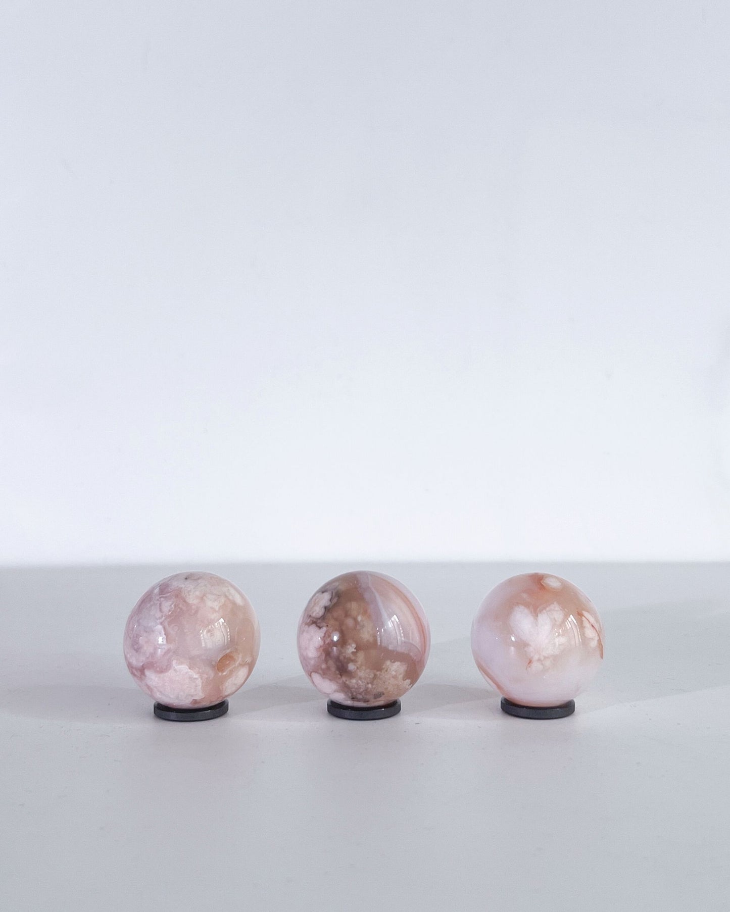 Three Carnelian Flower Agate spheres displayed upright on black stands against a minimal white background. Each piece shows varying patterns of pink, cream, and soft orange inclusions.