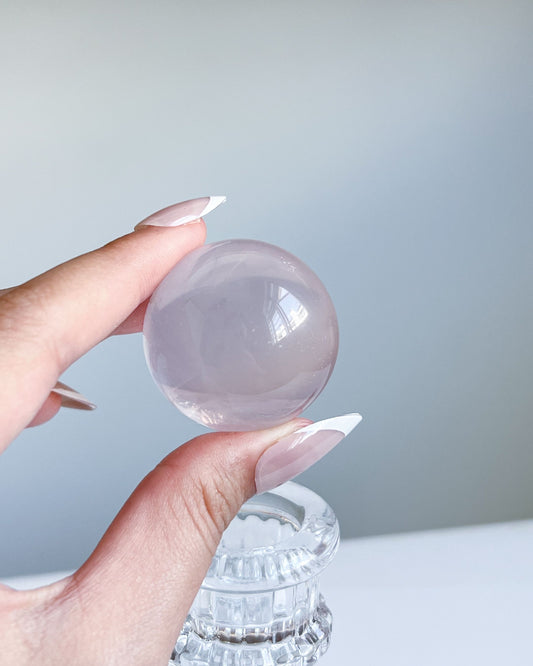 Hand holding a soft pink Girasol Rose Quartz sphere against a light grey background, with the crystal resting atop a clear ribbed glass stand