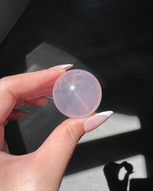 Girasol Rose Quartz sphere held under direct sunlight, showing glowing internal veils and a soft internal starburst reflection, set against a dark background.