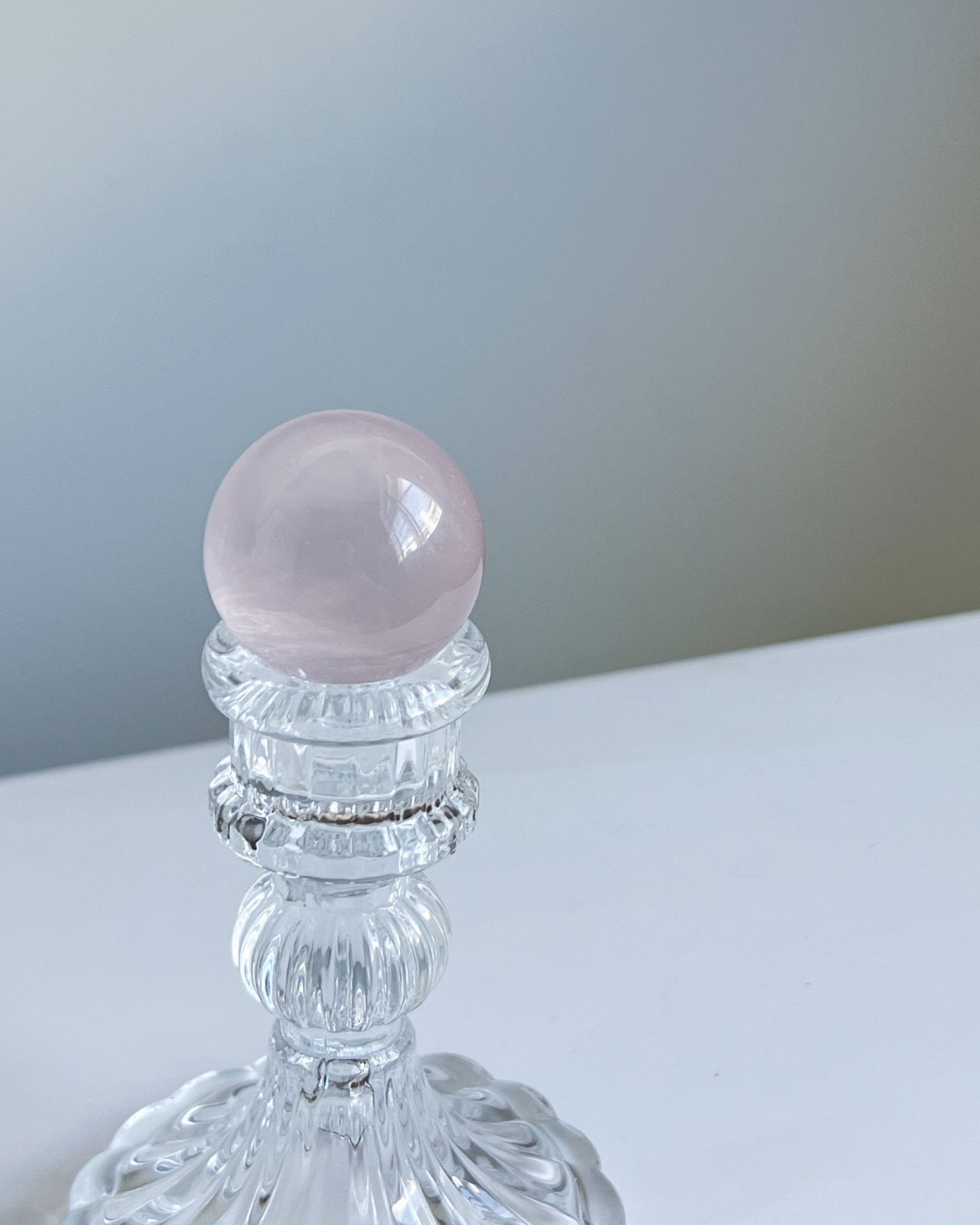 Girasol Rose Quartz sphere resting on a clear glass holder, with a soft neutral backdrop and even lighting that highlights the crystal’s blush tone.
