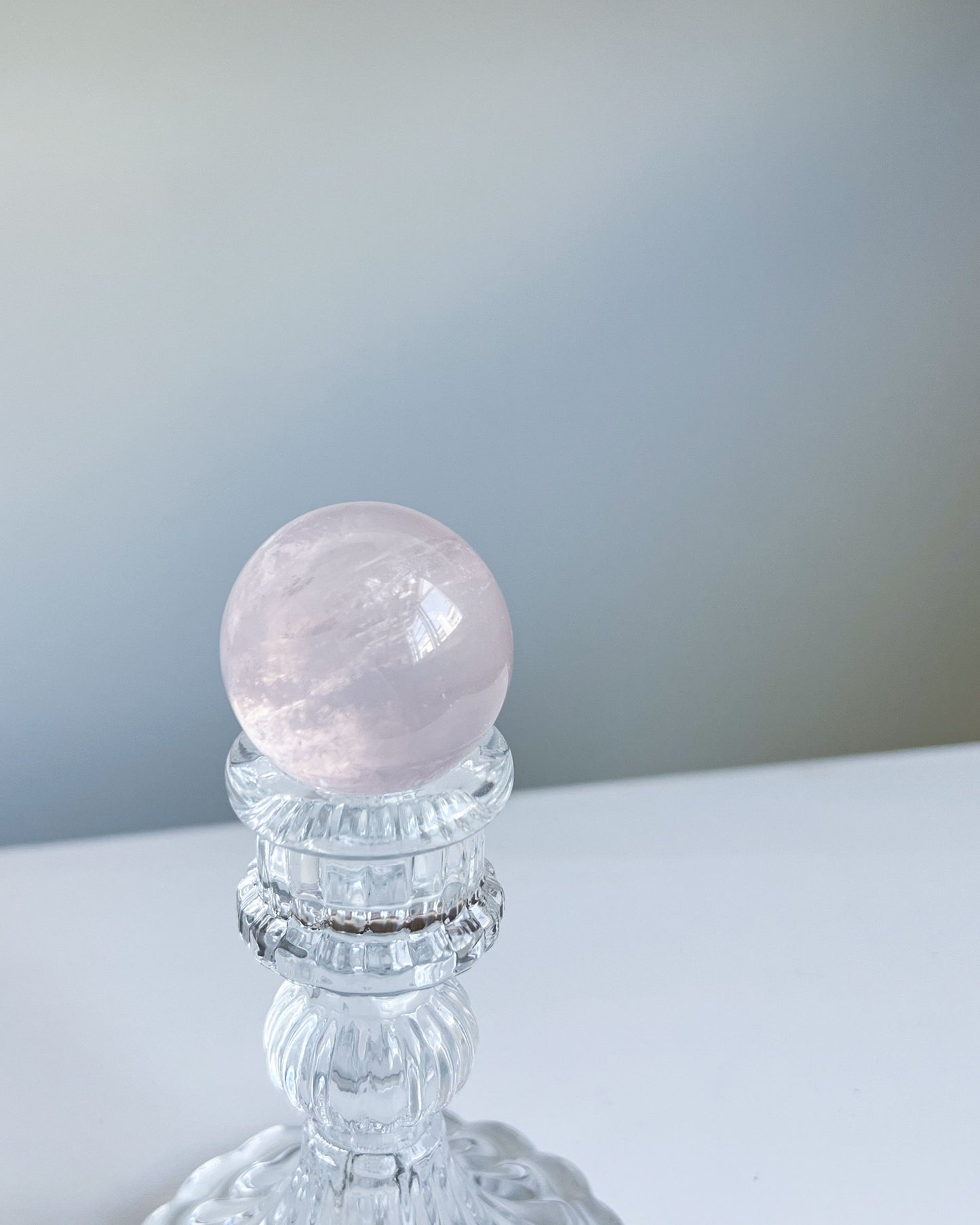 Polished Girasol Rose Quartz crystal sphere displayed on a clear vintage-style glass stand, with soft natural light highlighting its inner glow.