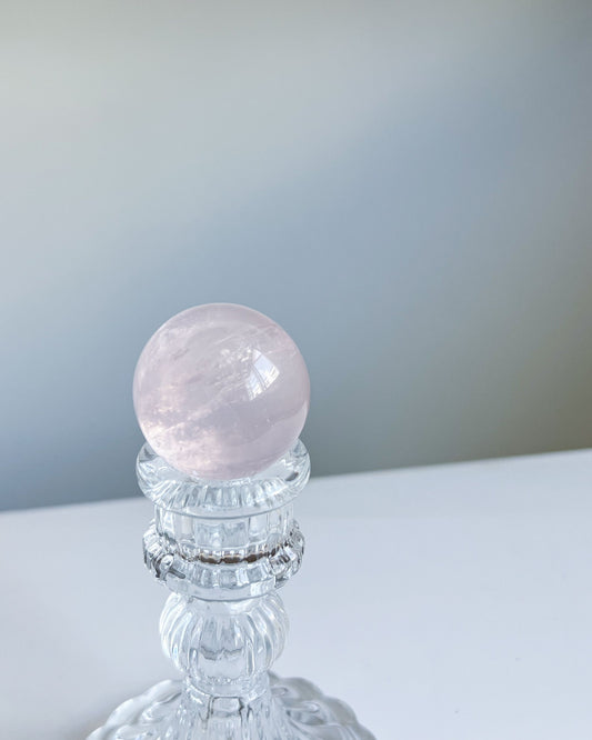 Polished Girasol Rose Quartz crystal sphere displayed on a clear vintage-style glass stand, with soft natural light highlighting its inner glow.