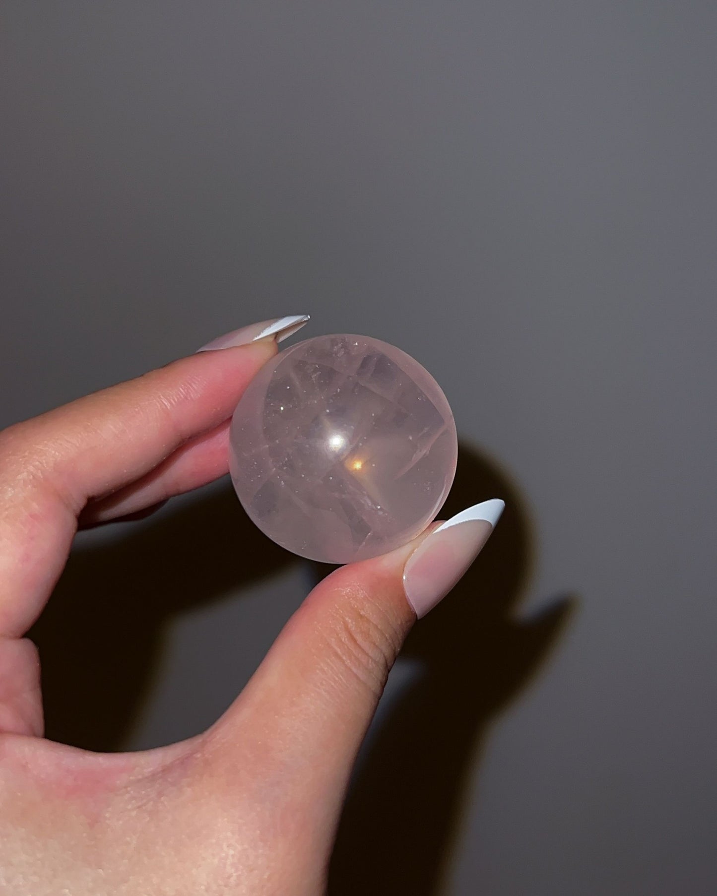 Girasol Rose Quartz gemstone sphere glowing under flash lighting, with internal striations and golden-orange glow against a dark grey wall.