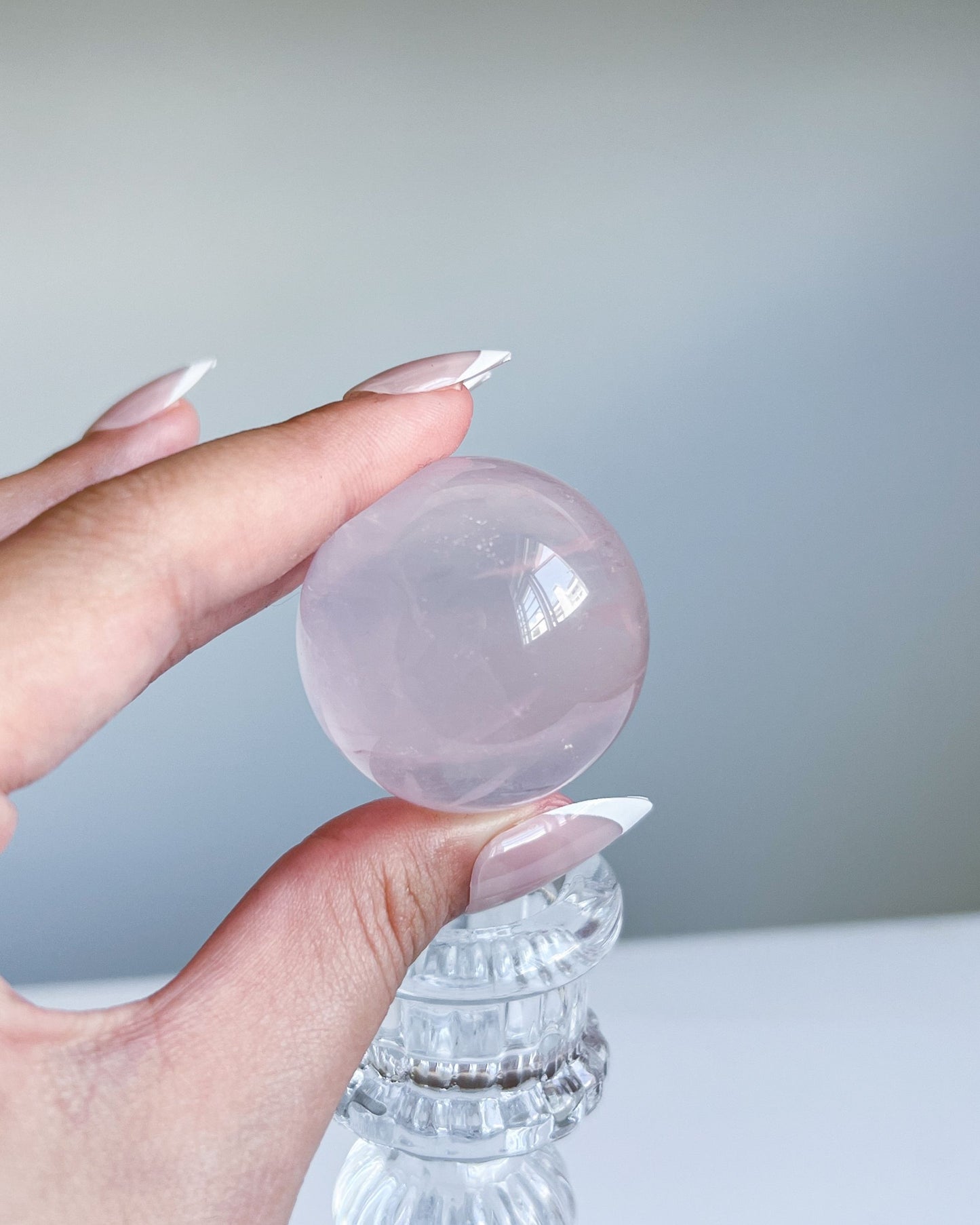 Hand holding Girasol Rose Quartz sphere atop a clear candlestick base, showcasing the blush pink hue and natural patterns of the crystal.