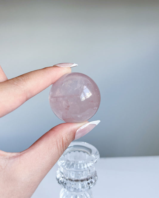 Girasol Rose Quartz crystal sphere held above a clear glass stand, featuring delicate pink hues, natural veils, and a glossy finish.