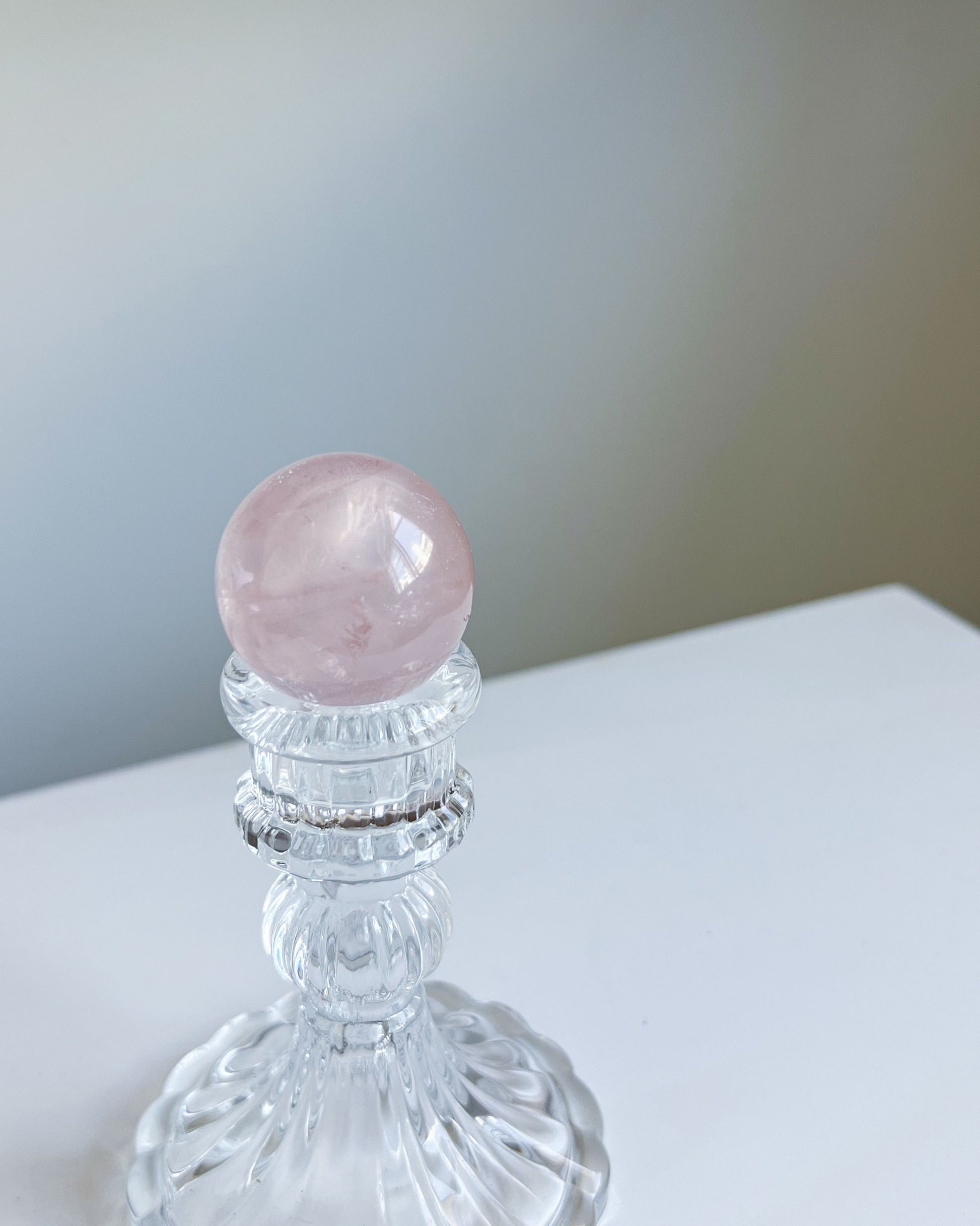 Girasol Rose Quartz crystal ball displayed on a scalloped clear pedestal, softly lit to highlight its pastel blush tone and gentle inner clarity.
