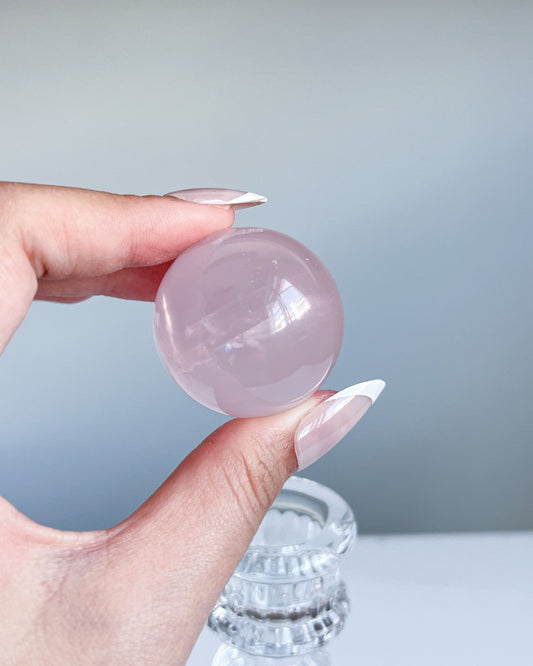 Girasol Rose Quartz crystal sphere held above a glass stand, with a glossy surface and delicate blush-pink tones reflecting soft daylight.