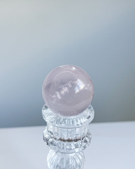 Girasol rose quartz sphere on glass stand under natural daylight, showcasing a dreamy soft pink glow and semi-translucent texture.