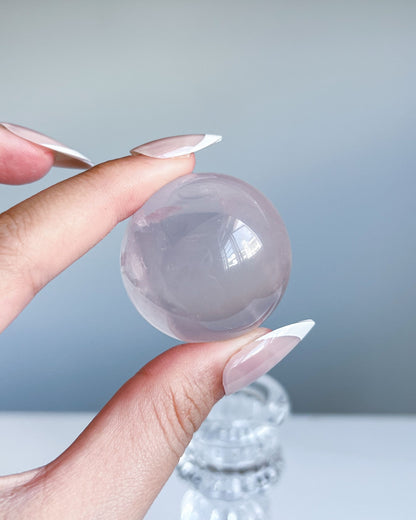 Light pink girasol rose quartz sphere held against a grey background, with soft inner whirls and glassy finish.