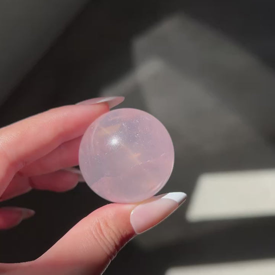 Close-up video of a Girasol Rose Quartz sphere held in hand in sunlight, with flash and studio lighting: revealing amazing asterism and ethereal glow.