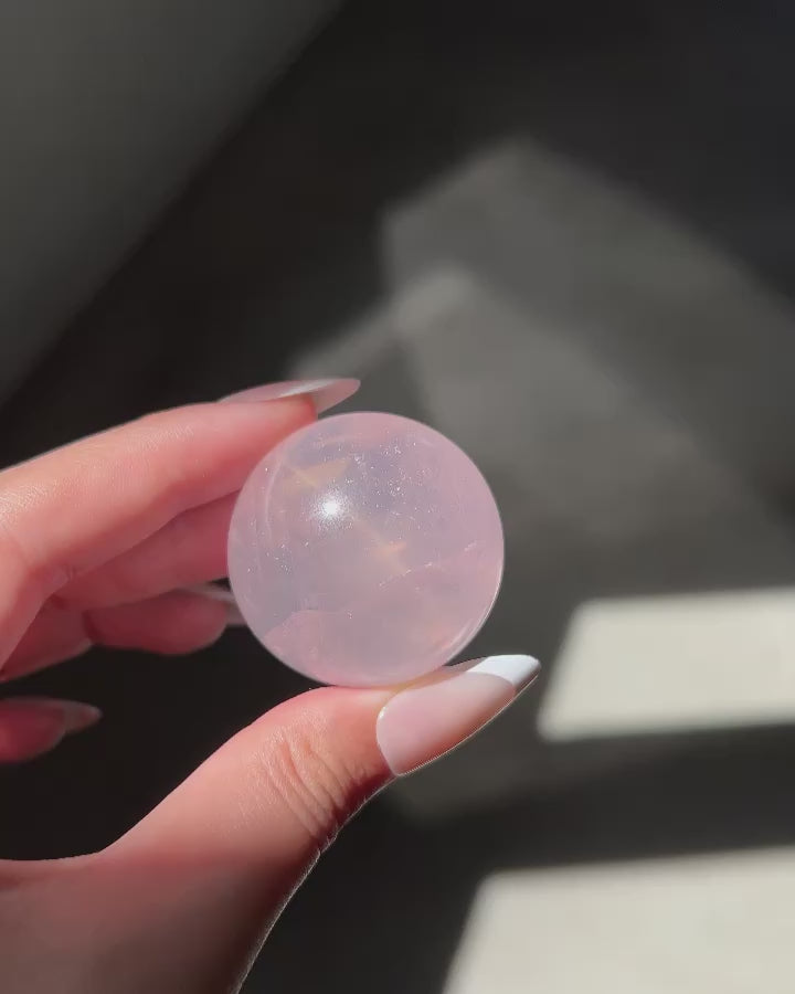 Close-up video of a Girasol Rose Quartz sphere held in hand in sunlight, with flash and studio lighting: revealing amazing asterism and ethereal glow.
